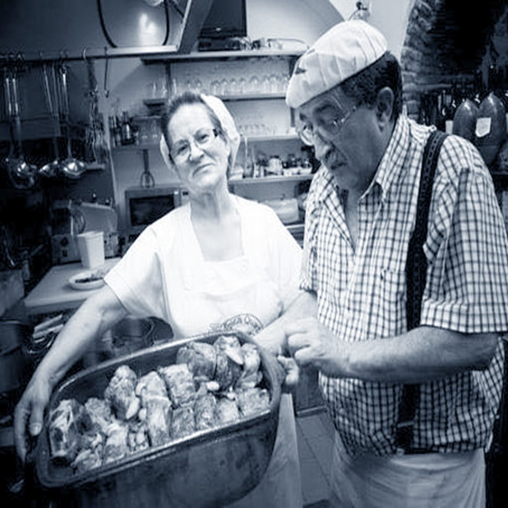 Foto a preto e branco numa taberna típica, com senhora cozinheira e homem segurando um tacho