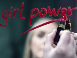 Texto "girl power" escrito pela mão de uma rapariga loura, num espelho, a batom vermelho